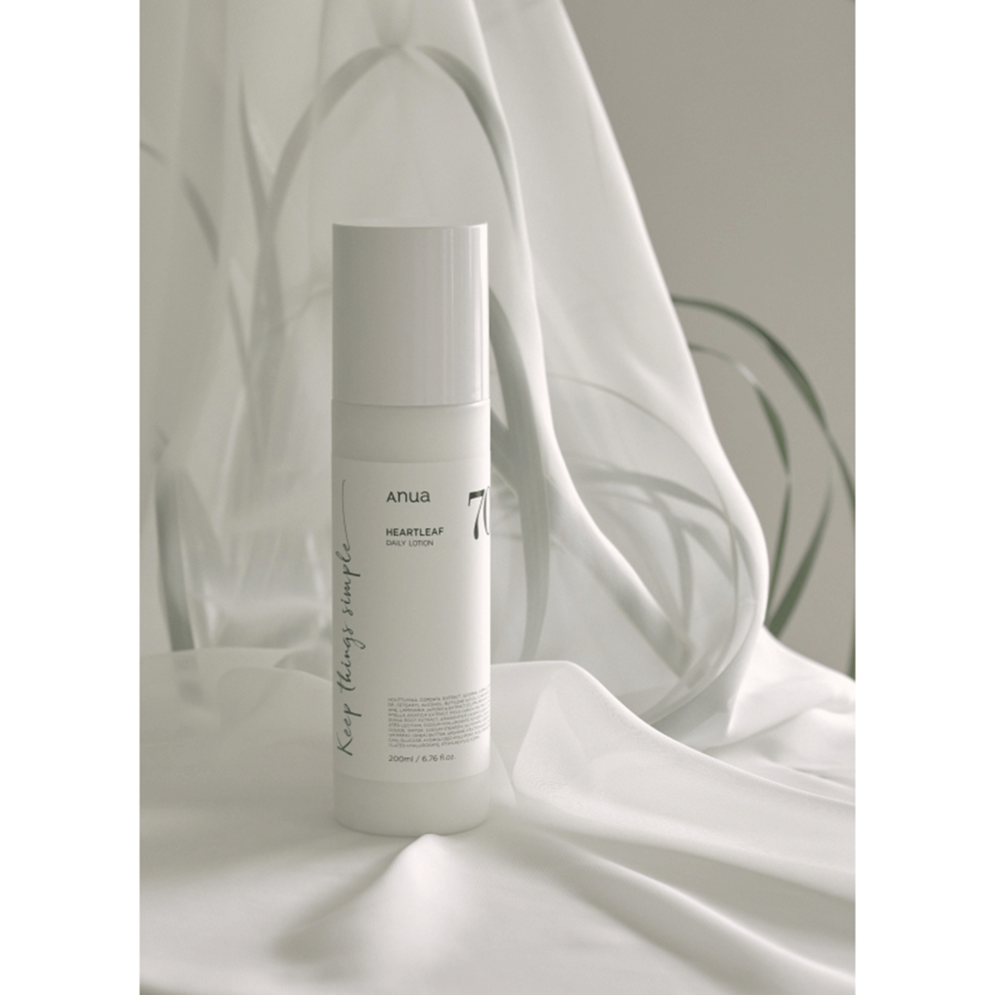 Eine weiße Flasche Anua Heartleaf 70% Daily Lotion 200ml steht aufrecht auf einem drapierten weißen Stoff, sanft beleuchtet mit verschwommenen Pflanzenschatten im Hintergrund.