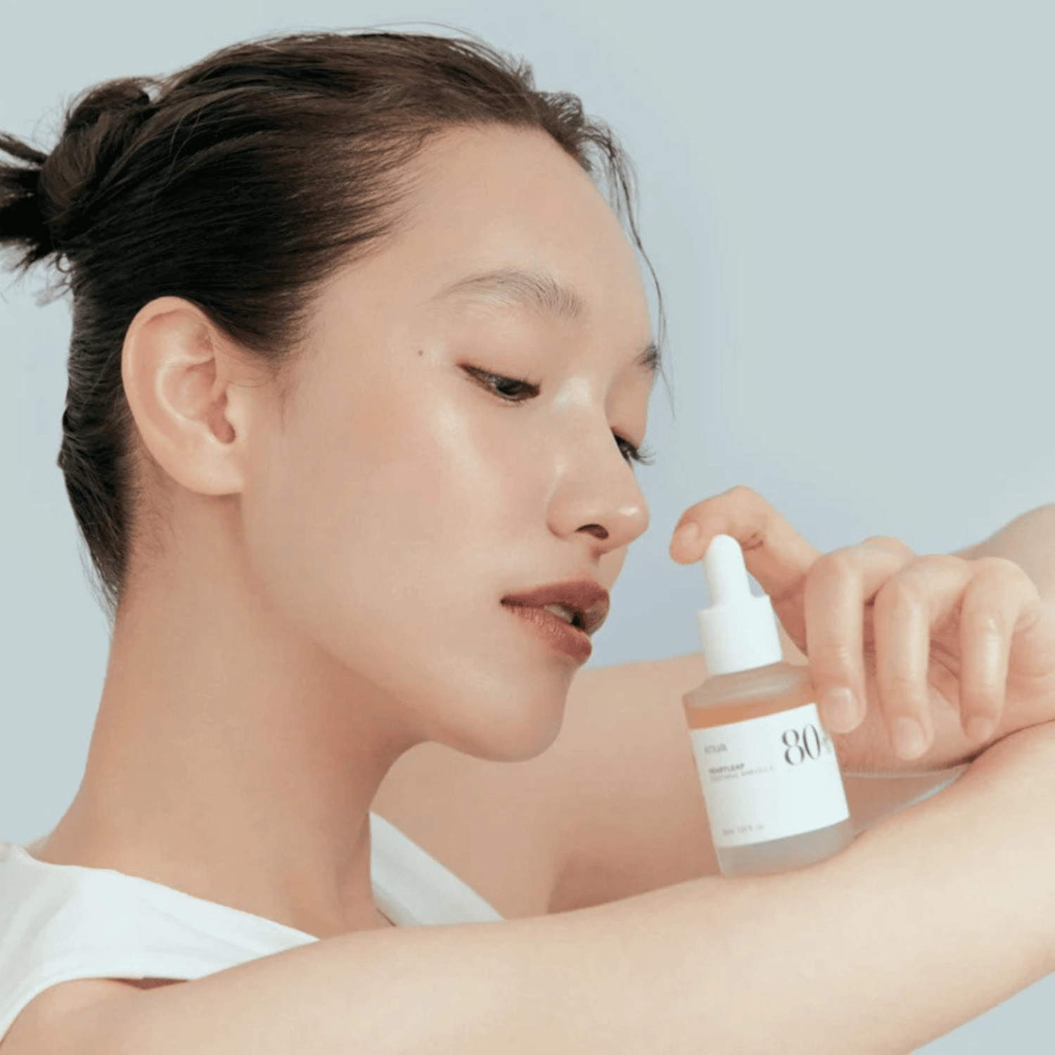 Eine Frau mit glatter Haut berührt sanft ihren Arm, während sie eine Flasche Anua Heartleaf 80% Soothing Ampoule 30ml in der Nähe ihres Gesichts hält, vor einem schlichten hellen Hintergrund.