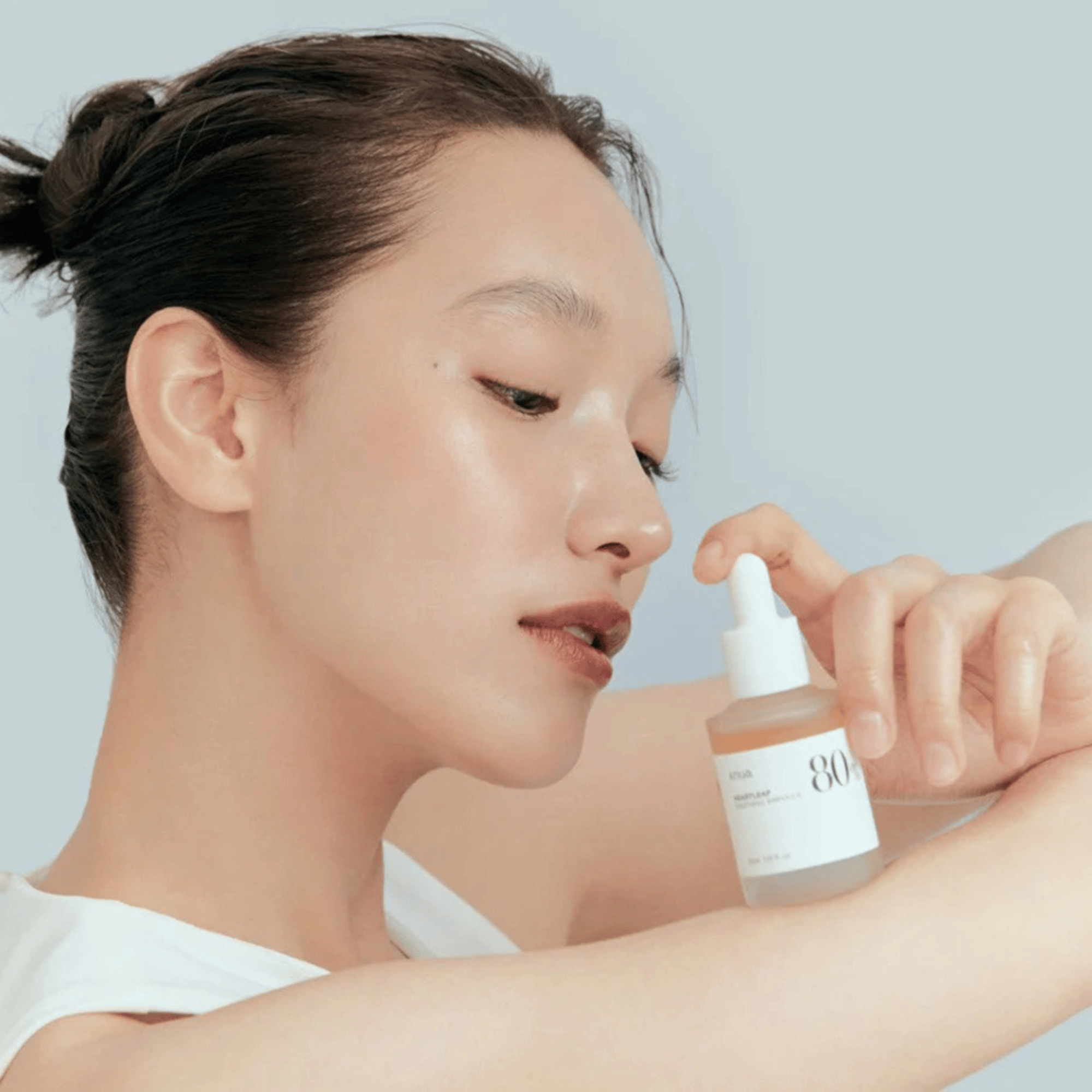 Eine Frau mit glatter Haut berührt sanft ihren Arm, während sie eine Flasche Anua Heartleaf 80% Soothing Ampoule 30ml in der Nähe ihres Gesichts hält, vor einem schlichten hellen Hintergrund.