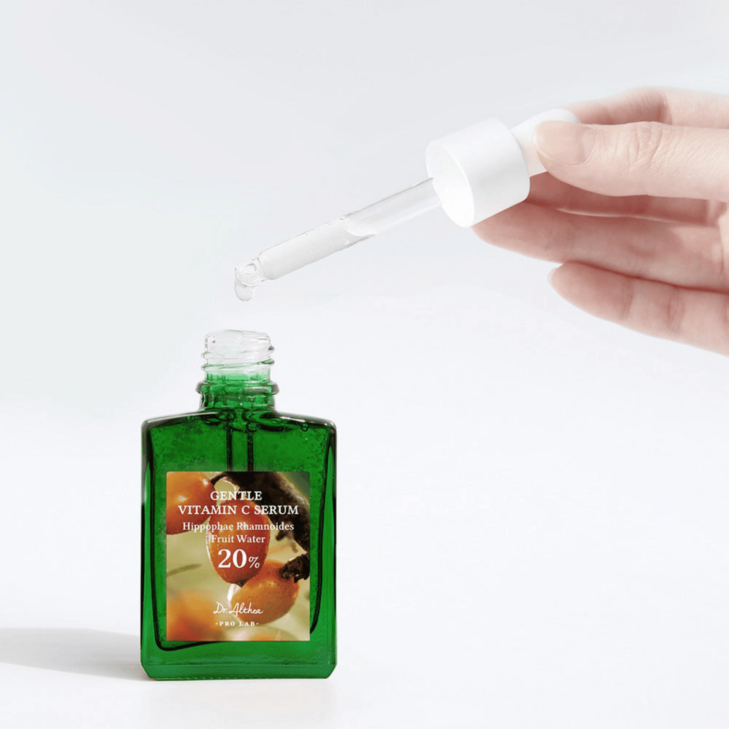 Eine Hand dosiert eine Pipette über eine grüne Glasflasche auf leicht grauen Hintergund.