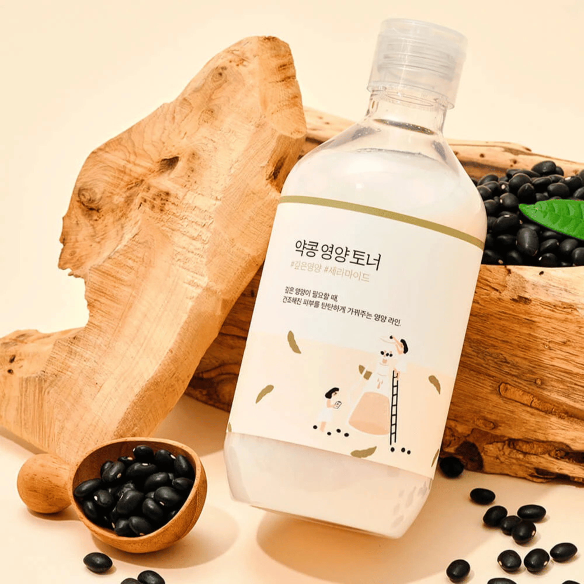 Die 300-ml-Flasche ROUND LAB Soybean Toner mit koreanischem Etikett steht neben einer Holzschüssel mit verstreuten schwarzen Bohnen und einem Holzscheit auf einer beigen Fläche.