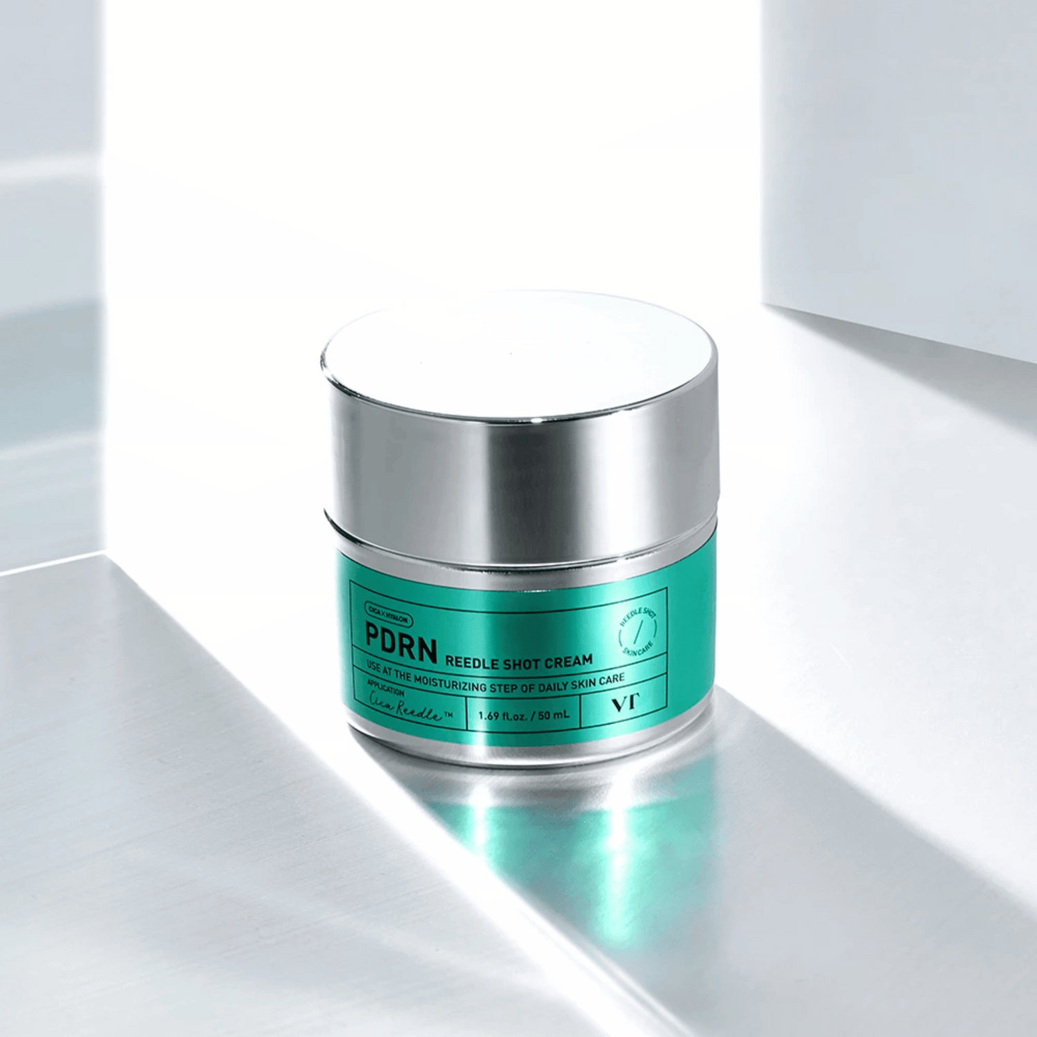 Ein silberner und türkisfarbener Tiegel mit der Aufschrift VT Cosmetics PDRN Reedle Shot Cream 50ml steht auf einer reflektierenden, minimalistischen Oberfläche mit sanfter Beleuchtung.