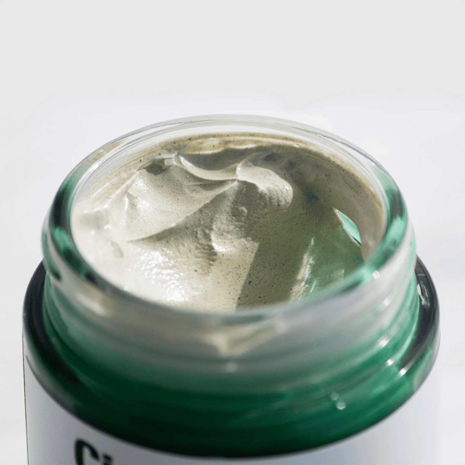 Ein geöffnetes Glas Dr. Jart+ Cicapair Tiger Grass Color Correcting Treatment mit seiner glatten, hellgrünen Creme in einem grünen Glasbehälter vor einem schlichten weißen Hintergrund.