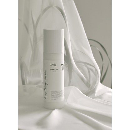 Eine weiße Flasche Anua Heartleaf 70% Daily Lotion 200ml steht aufrecht auf einem drapierten weißen Stoff, sanft beleuchtet mit verschwommenen Pflanzenschatten im Hintergrund.