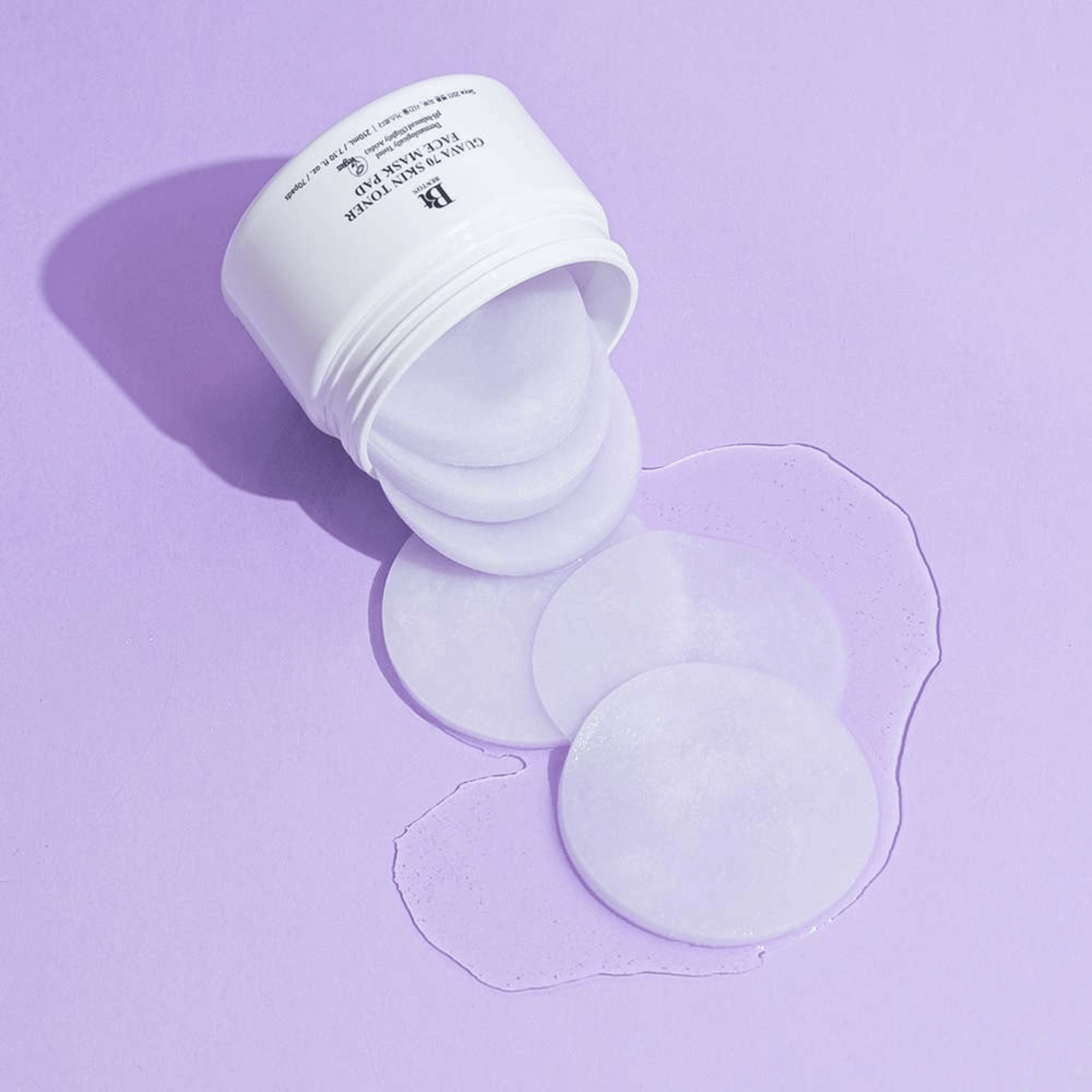 Ein weißer Behälter mit Benton Guava Skin Toner Face Mask Pads ist auf einer hellvioletten Oberfläche umgekippt, wobei etwas Flüssigkeit um die Pads herum verschüttet ist.