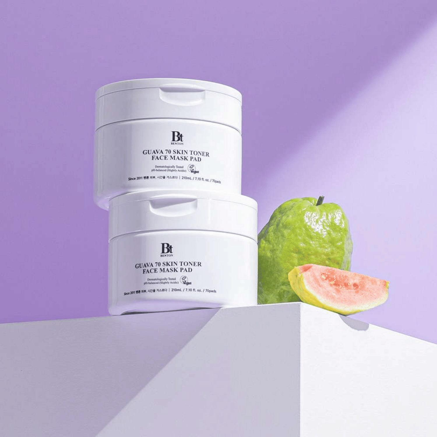 Zwei Benton Guava Skin Toner Face Mask Pad sind neben einer ganzen Guave und einer Scheibe auf einer weißen Fläche gestapelt, alles vor einem hellvioletten Hintergrund.
