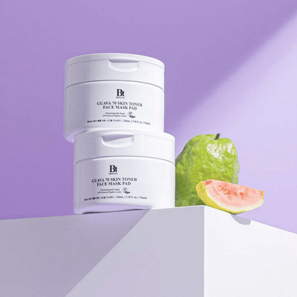 Zwei Benton Guava Skin Toner Face Mask Pad sind neben einer ganzen Guave und einer Scheibe auf einer weißen Fläche gestapelt, alles vor einem hellvioletten Hintergrund.